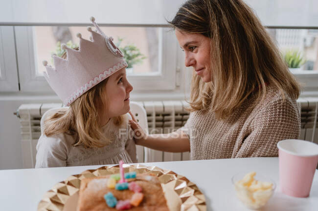 Vista lateral da mãe alegre colocando sentiu coroa artesanal na filha ao celebrar o aniversário juntos em casa — Fotografia de Stock