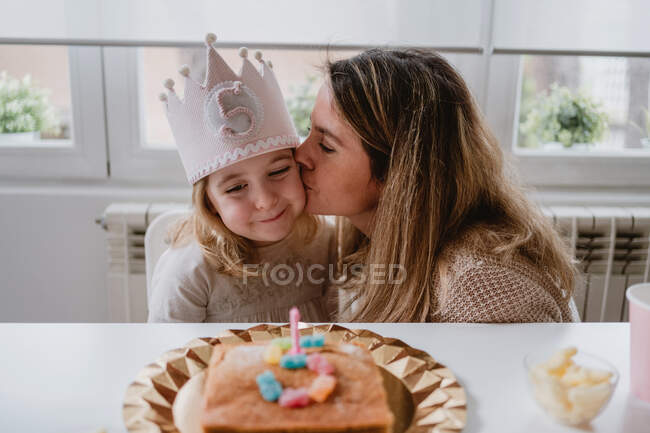 Madre cariñosa besando y felicitando a la niña pequeña juntos mientras pasan tiempo durante la fiesta de cumpleaños en la mesa en casa - foto de stock