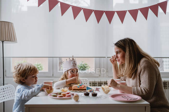 Madre positiva in abiti casual seduta al tavolo di legno e che parla con i bambini che festeggiano il compleanno a casa — Foto stock