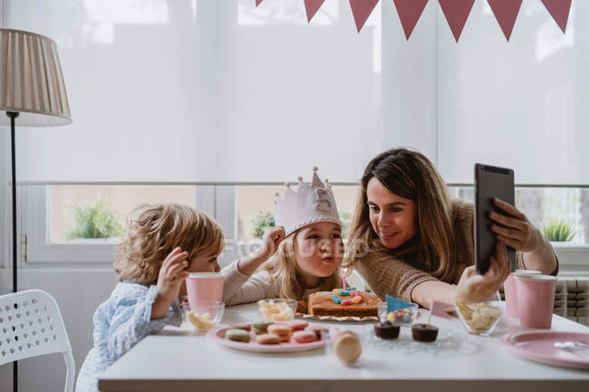 Mãe e filha positivas em roupas casuais sentadas juntas à mesa e fazendo videochamada no tablet enquanto comemoram o aniversário em casa — Fotografia de Stock