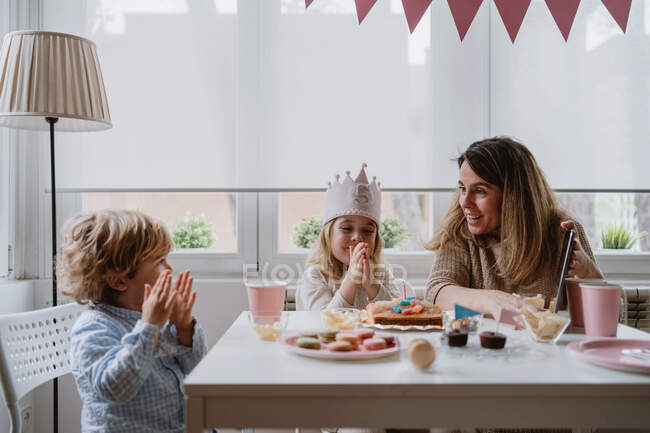 Madre e figlia positive in abiti casual seduti insieme a tavola e fare videochiamate sul tablet mentre festeggiano il compleanno a casa — Foto stock