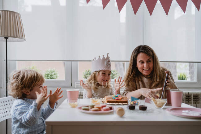 Madre e figlia positive in abiti casual seduti insieme a tavola e fare videochiamate sul tablet mentre festeggiano il compleanno a casa — Foto stock
