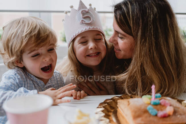 Madre cariñosa y hermano felicitando a la niña pequeña juntos mientras pasan tiempo durante la fiesta de cumpleaños en la mesa en casa - foto de stock