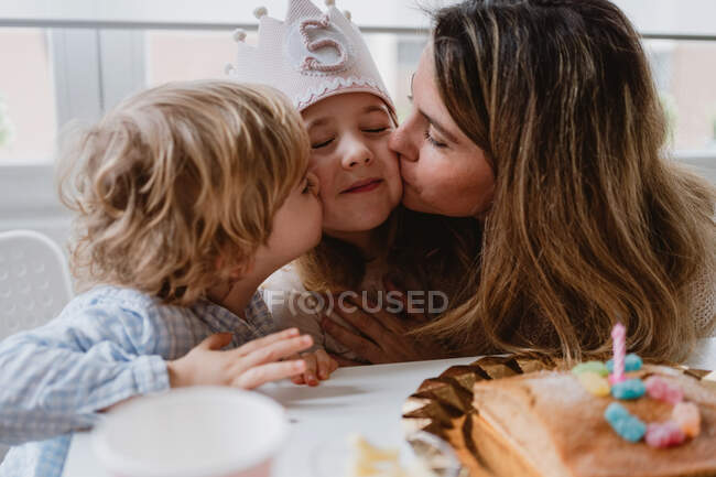 Madre y hermano cariñosos besando y felicitando a la niña mientras pasan tiempo juntos durante la fiesta de cumpleaños en la mesa en casa - foto de stock