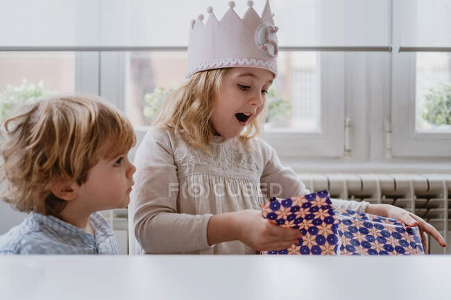 Emocionado chica encantadora en la corona hecha a mano desenvolver caja de regalo mientras que tiene celebración de cumpleaños en casa - foto de stock