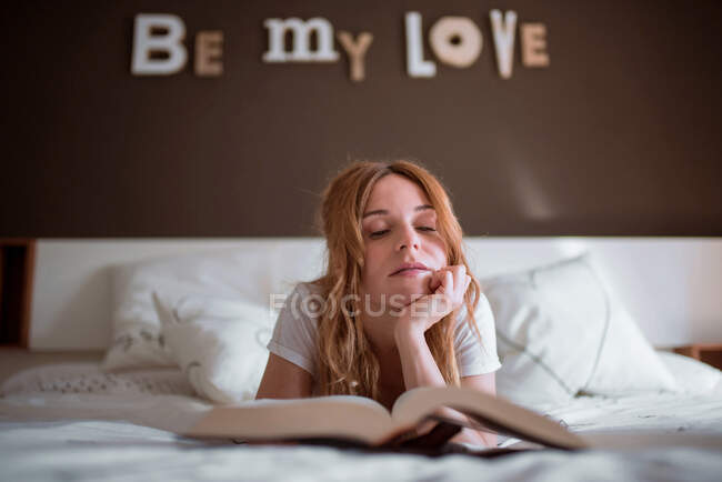 Мирна жінка відпочиває на ліжку, лежачи на животі і читаючи цікаву книжку в затишній спальні з романтичним написом на стіні і мрійливою атмосферою — стокове фото
