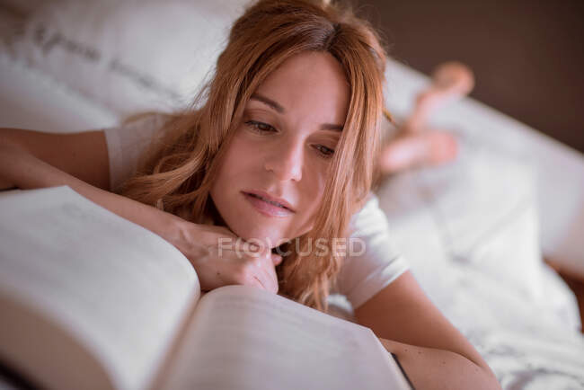 Femme paisible se détendre sur le lit tout en étant couché sur le ventre et la lecture livre intéressant dans une chambre confortable avec inscription romantique sur le mur et atmosphère rêveuse — Photo de stock