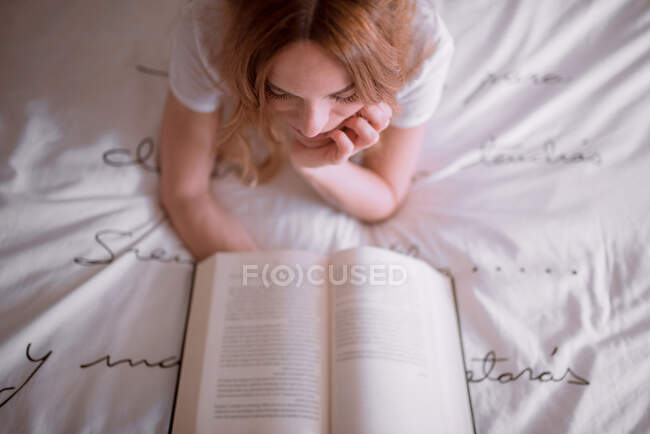 Мирная женщина отдыхает на кровати, лежа на животе и читая интересную книгу в уютной спальне с романтической надписью на стене и мечтательной атмосферой — стоковое фото