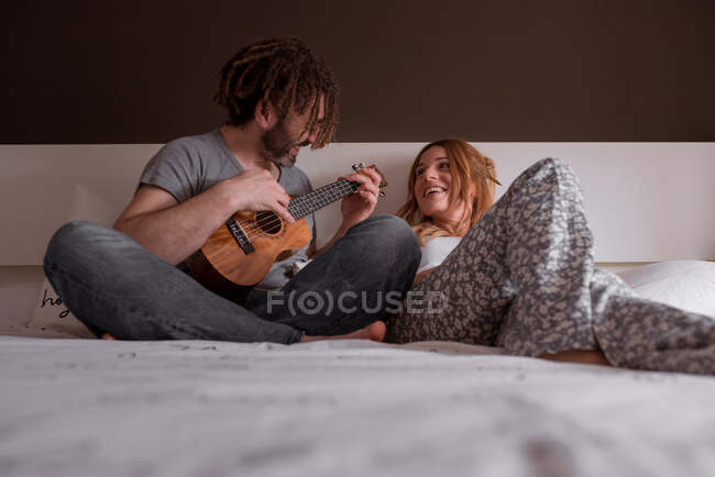 Alegre joven con rastas y mujer con el pelo rojo sentado en la cama y divertirse con los ojos cerrados tocando la guitarra ukelele mientras pasan tiempo juntos en casa en fin de semana - foto de stock