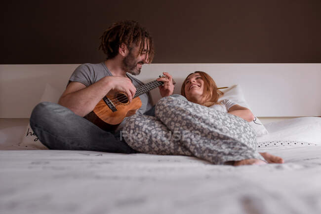 Cheerful young man with dreadlocks and woman with red hair sitting on bed and having fun with closed eyes playing ukulele guitar while spending time together at home at weekend — Stock Photo