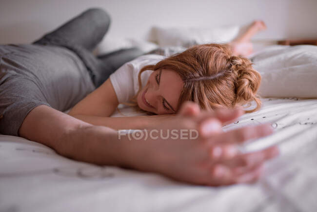 Hombre de las cosechas abrazar y besar a la mujer con el pelo rojo mientras yacen en el estómago en la cama juntos y relajarse en fin de semana - foto de stock