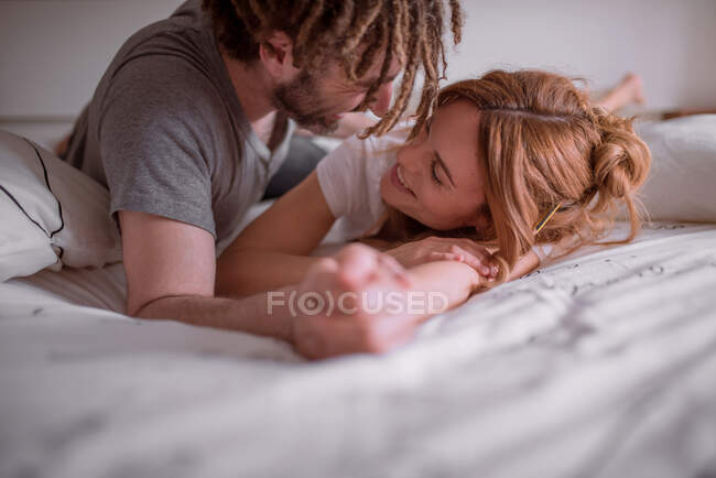Zarter Mann mit Dreadlocks umarmt und küsst Frau mit roten Haaren, während er auf dem Bauch zusammen im Bett liegt und sich am Wochenende entspannt — Stockfoto