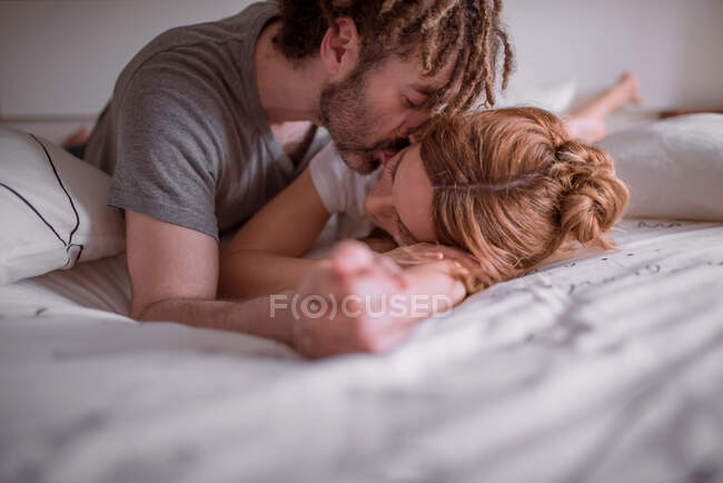 Tenero uomo con dreadlocks abbracciare e baciare donna con i capelli rossi mentre sdraiati sullo stomaco sul letto insieme e rilassante nel fine settimana — Foto stock