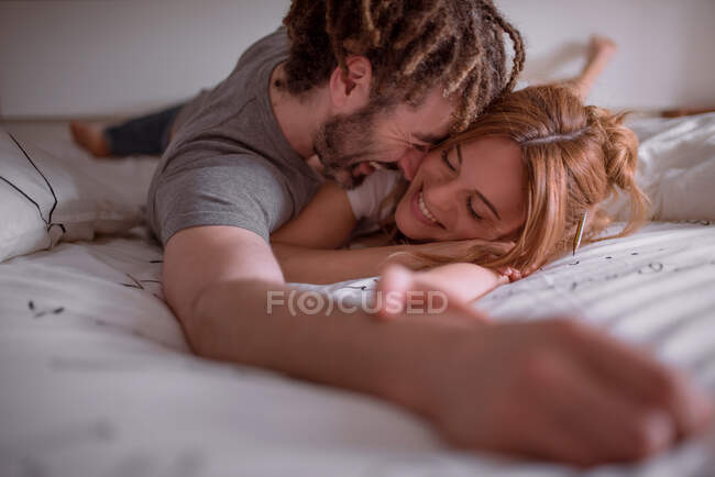 Ніжний чоловік з дредлоками обіймає і цілує жінку червоним волоссям, лежачи на шлунку на ліжку разом і розслабляючись у вихідні — стокове фото
