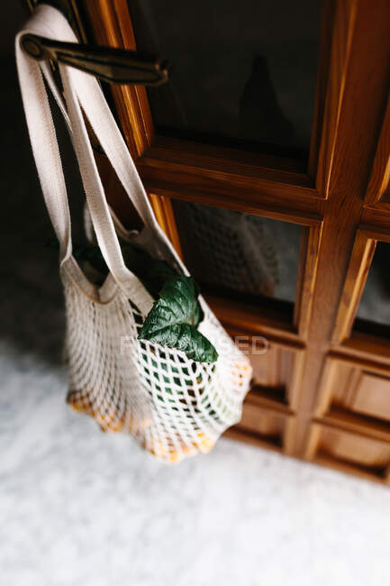 Біла нитяна сумка зі свіжими пухкими фруктами і листям, що звисають на дверях — стокове фото