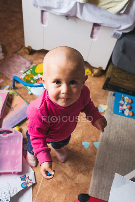 Dall'alto vista di carino bambino in camicia rosa casual in piedi sul pavimento in sala giochi leggera e guardando la fotocamera mentre trascorre del tempo a casa — Foto stock