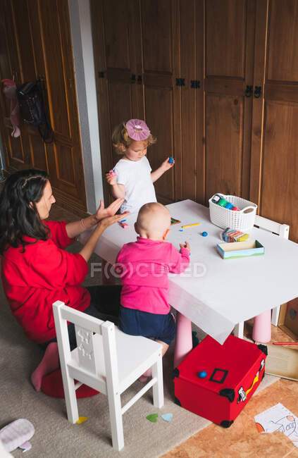Позитивна мати в повсякденному червоному одязі сидить за столом з дітьми і грає з пластиліном, проводячи час вдома — стокове фото