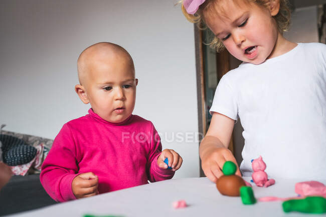 Petits enfants joyeux et mignons en vêtements décontractés jouant avec la plasticine tout en passant du temps ensemble à la maison — Photo de stock