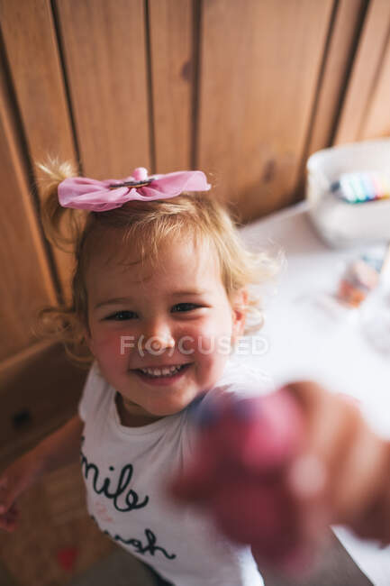 Усміхнена маленька дівчинка в білій повсякденній сорочці тримає іграшку з пластмаси ручної роботи і дістається до камери, граючи вдома — стокове фото
