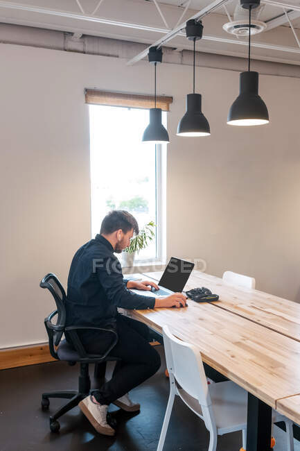 Empresario masculino sentado en la mesa de madera en un espacio de trabajo creativo y trabajando en un proyecto remoto mientras usa netbook - foto de stock