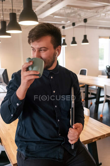 Alegre freelancer masculino en auriculares inalámbricos y con portátil apoyado en la mesa en un espacio de trabajo creativo y disfrutando de una bebida caliente mientras toma un descanso durante el trabajo remoto - foto de stock