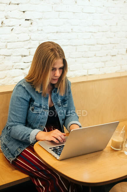 Alto ángulo de mujer autónoma concentrada en ropa casual sentada a la mesa en un espacio de trabajo brillante y escribiendo en netbook durante el trabajo remoto - foto de stock