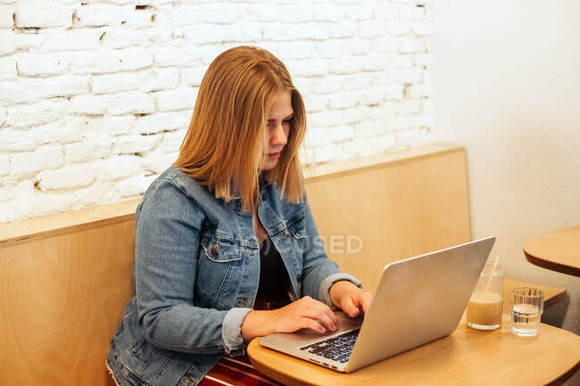 High angle of concentrated self employed female in casual clothes sitting at table in bright workspace and typing on netbook during remote work — Stock Photo