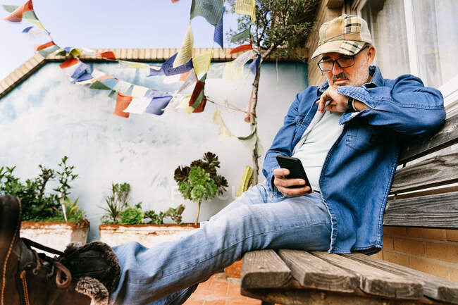 Ruhige nachdenkliche Männchen mit Brille und Mütze, die sich auf Bildschirm und Nachrichten mit dem Smartphone konzentrieren, während sie es sich auf einer Holzbank im Hinterhof eines Ferienhauses in einem Vorort gemütlich machen — Stockfoto