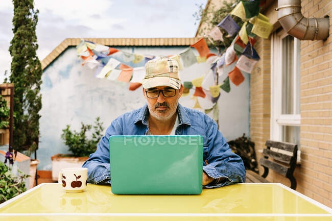Задумчивый пожилой мужчина в шапочке и джинсовой рубашке с интересом сидит за столом с кружкой вкусного напитка и использует нетбук во дворе загородного дома — стоковое фото