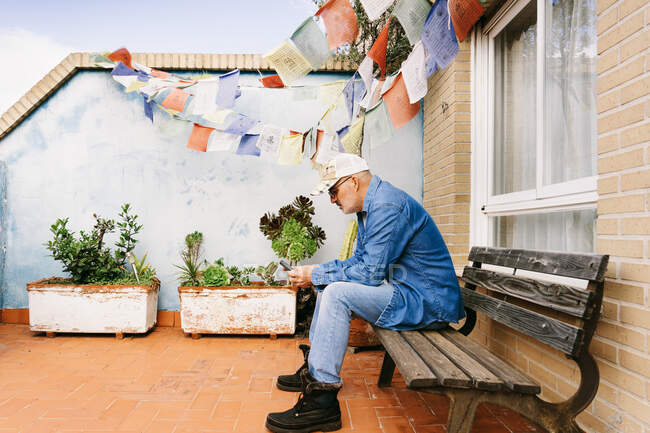 Продуманий сіро-волохатий чоловік в джинсовому одязі, використовуючи смартфон, коли він охолоджується в саду — стокове фото