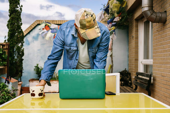 Задумчивый пожилой мужчина в шапочке и джинсовой рубашке с интересом сидит за столом с кружкой вкусного напитка и использует нетбук во дворе загородного дома — стоковое фото