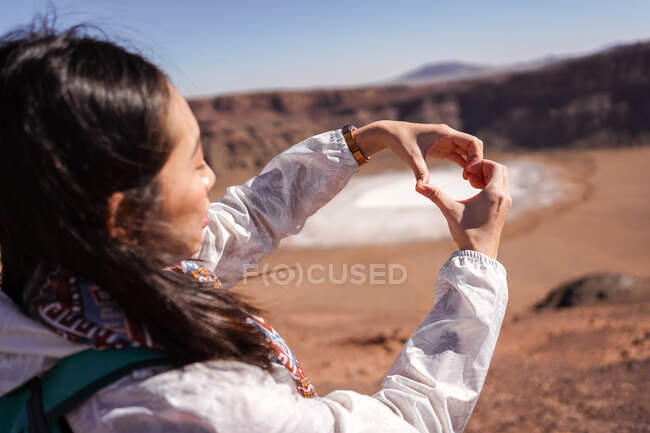 Inspirada viajera asiática vistiendo ropa casual y mochila haciendo corazón con los dedos mientras está de pie contra el cráter de maar en el desierto con terreno rocoso - foto de stock