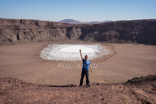 Удаленное представление взволнованного путешественника-мужчины в повседневной одежде и солнечных очках, указывающих вверх с поднятой рукой, отмечающих направление живописной скалистой местности с белой поверхностью фосфата натрия внутри кратера — стоковое фото
