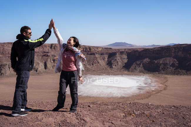 Jovem homem e mulher multirraciais excitados dando altos cinco enquanto viajam juntos para a cratera maar com superfície de cristal de fosfato de sódio branco — Fotografia de Stock