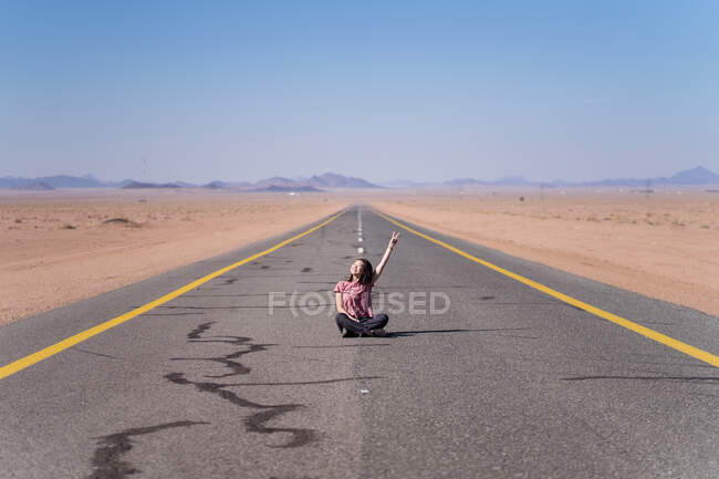 Sonriente adolescente en ropa casual sentado con las piernas cruzadas en el camino de asfalto con marcado amarillo y blanco cerca de la arena y las montañas detrás y demostrando gesto de paz con la mano levantada - foto de stock