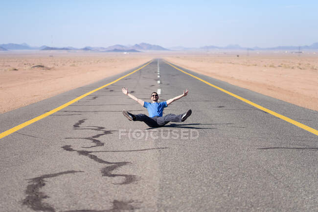 Hombre sonriente en ropa casual sentado con las piernas cruzadas en el camino de asfalto cerca de la arena y las montañas detrás y demostrando gesto de paz con la mano levantada - foto de stock