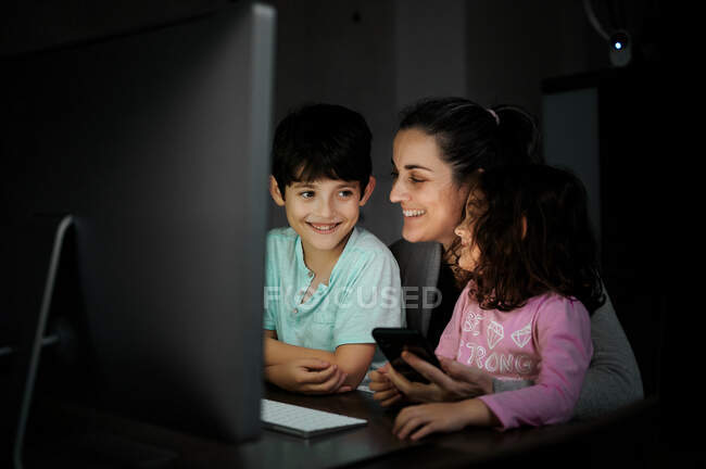 Jovem mãe feliz com filho e filha segurando smartphone enquanto sentados juntos na mesa com o computador e assistindo vídeo engraçado no quarto escuro em casa — Fotografia de Stock