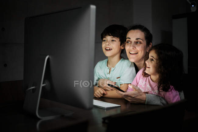 Счастливая молодая мать с маленьким сыном и дочерью держат смартфон, сидя вместе за столом с компьютером и смотреть смешное видео в темной комнате дома — стоковое фото