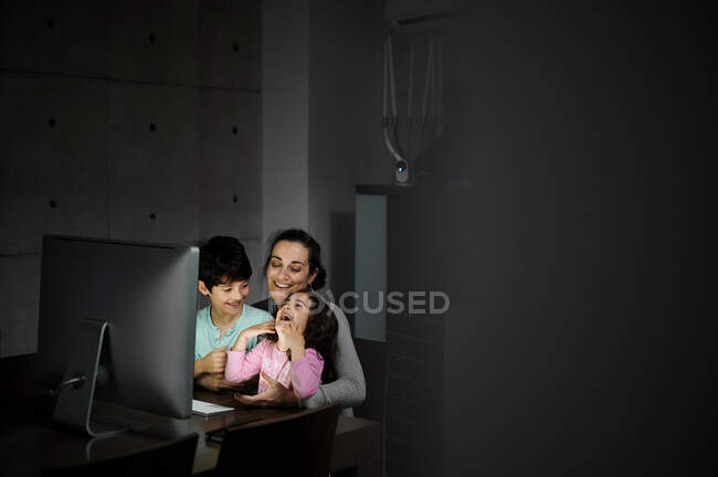 Glückliche junge Mutter mit kleinem Sohn und Tochter, die das Smartphone in der Hand halten, während sie zusammen am Tisch mit dem Computer sitzen und zu Hause lustige Videos im dunklen Raum ansehen — Stockfoto