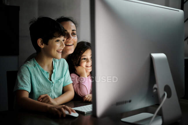Glückliche junge Mutter mit kleinem Sohn und Tochter, die das Smartphone in der Hand halten, während sie zusammen am Tisch mit dem Computer sitzen und zu Hause lustige Videos im dunklen Raum ansehen — Stockfoto