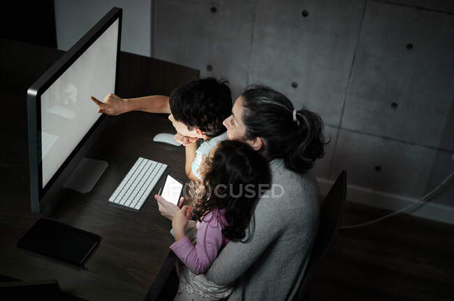 Happy young mother with little son and daughter holding smartphone while  sitting together at table with computer and watching funny video in dark  room at home — having fun, telecommuting - Stock