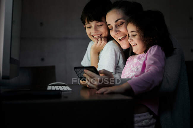 Felice giovane madre con piccolo figlio e figlia che tiene lo smartphone mentre si siede insieme a tavola con il computer e guarda video divertenti in camera oscura a casa — Foto stock