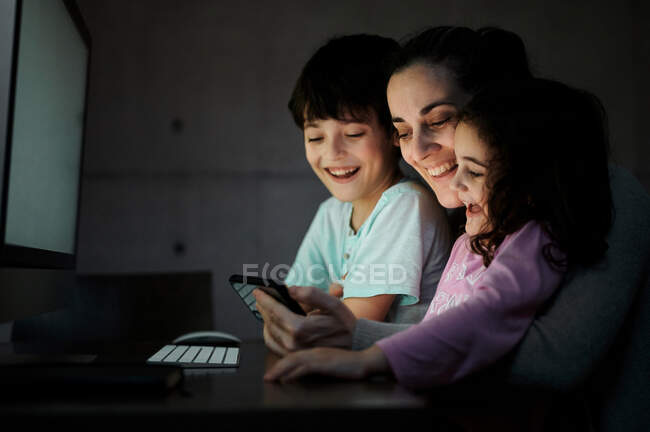 Joyeux jeune mère avec petit fils et fille tenant smartphone tout en étant assis ensemble à table avec ordinateur et en regardant une vidéo drôle dans la chambre noire à la maison — Photo de stock
