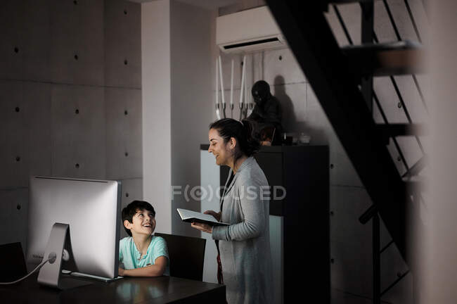 Вид сбоку молодой женщины, объясняющей учебное задание положительному сыну, сидящему за столом с компьютером и учебником во время онлайн-урока дома — стоковое фото