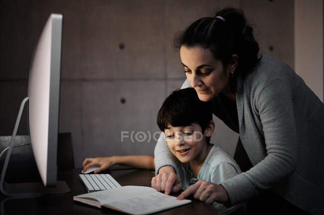 Vue latérale de la jeune femme expliquant la tâche d'étude au fils positif assis à table avec ordinateur et manuel pendant la leçon en ligne à la maison — Photo de stock