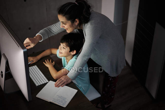 Vista lateral de la joven mujer explicando la tarea de estudio al hijo positivo sentado en la mesa con la computadora y el libro de texto durante la lección en línea en casa - foto de stock