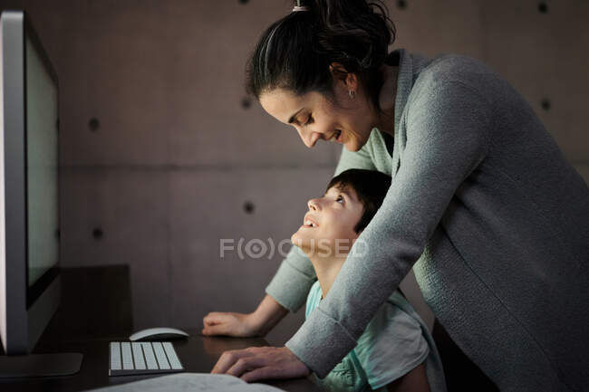 Side view of young woman explaining study task to positive son sitting at table with computer and textbook during online lesson at home — Stock Photo