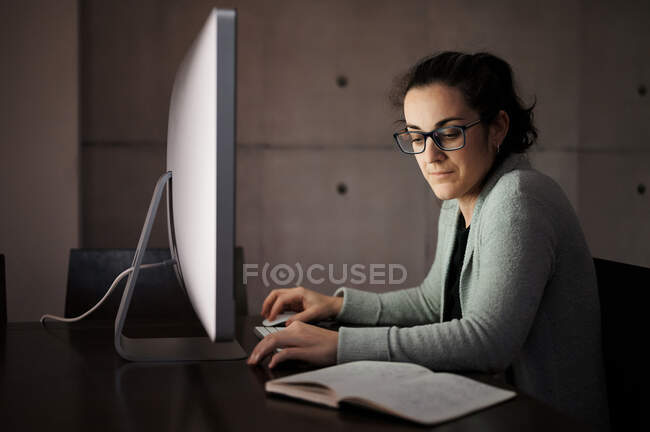 Visão lateral da fêmea jovem concentrada séria em desgaste casual e óculos sentados à mesa com computador com tela branca em branco e smartphone de navegação enquanto trabalhava remotamente à noite em casa — Fotografia de Stock