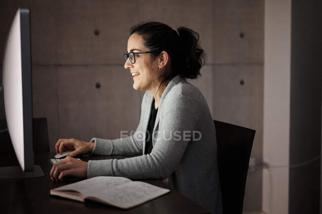 Вид збоку серйозної концентрованої молодої жінки в повсякденному одязі та окулярах, що сидять за столом з комп'ютером з білим порожнім екраном та переглядом смартфона, працюючи віддалено ввечері вдома — стокове фото