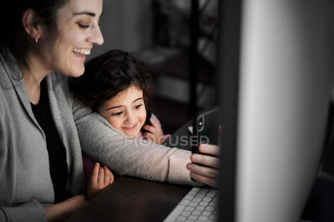 Joyeux jeune femme ethnique avec petite fille utilisant un téléphone portable et communiquant avec des amis par chat vidéo tout en passant du temps à la maison — Photo de stock